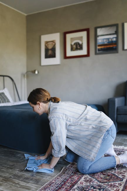 A person kneeling on a patterned carpet in a home setting, preparing a compact yellow and black vacuum cleaner for surface cleaning. The individual is wearing a beige jacket and blue jeans, with their