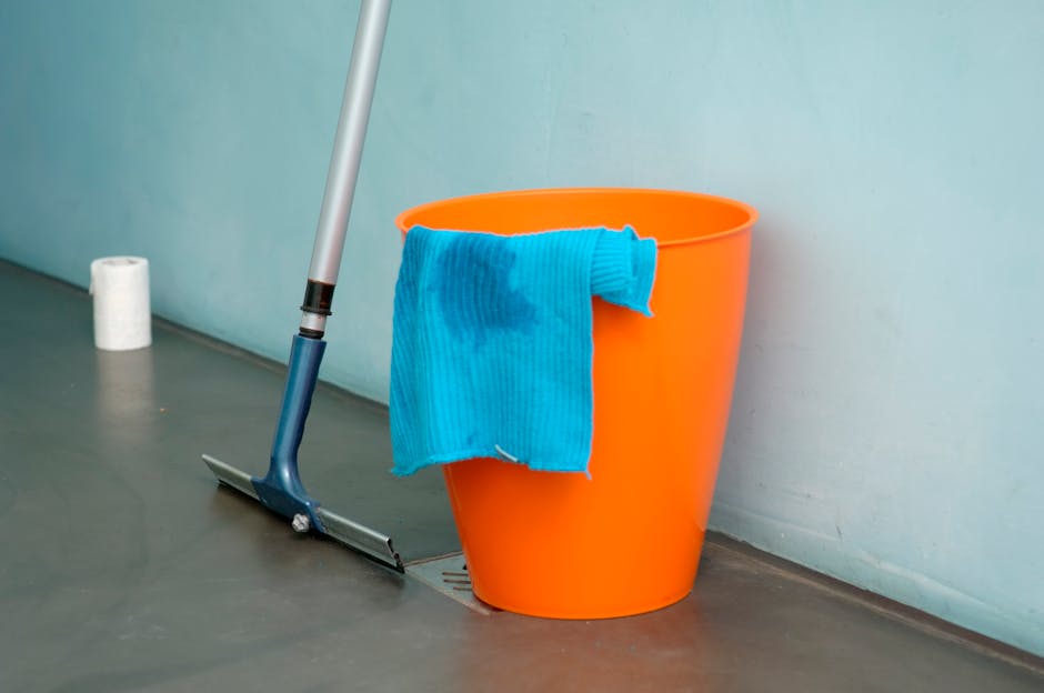 An indoor scene showing a bright orange plastic bucket partially covered with a blue microfiber cloth that is stained, placed on a smooth, dark gray floor next to a drain. A mop with a blue handle and